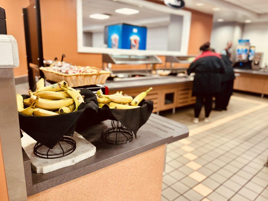 Cafeteria setting with a fruit station featuring bananas in the foreground. Two people are seen serving food from the buffet area in the background.