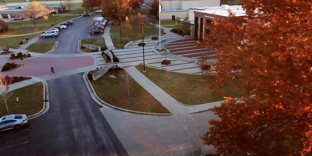 Aerial view of a modern building with a large parking lot, neatly landscaped areas, and scattered cars. The area is surrounded by autumn-colored trees.